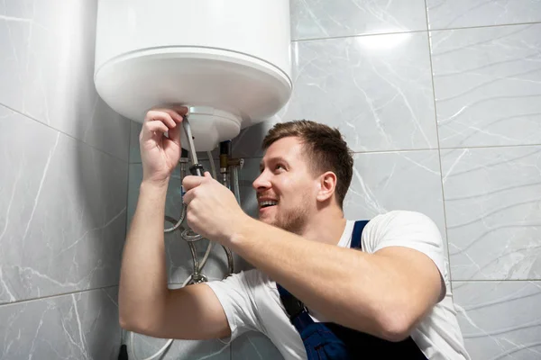 Joven guapo sonriente trabajador en uniforme de reparación de calentador de agua con destornillador en casa en el servicio de reparación profesional toilette — Foto de Stock