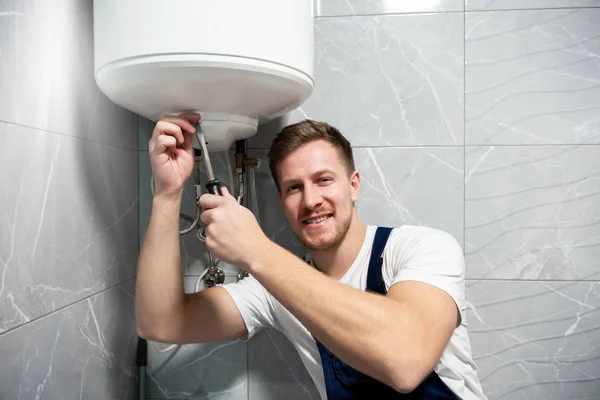 Joven guapo sonriente trabajador en uniforme con destornillador en la mano reparación de calentador de agua en casa en el servicio de reparación profesional toilette — Foto de Stock