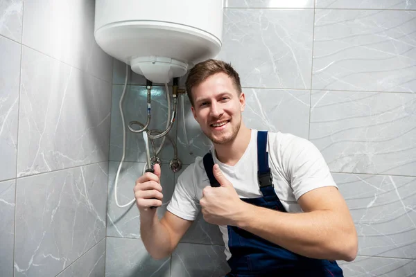 Sonriente hombre trabajador en uniforme con destornillador en una mano y mostrando como con otra mano reparación de caldera en casa en servicio de reparación profesional toilette — Foto de Stock