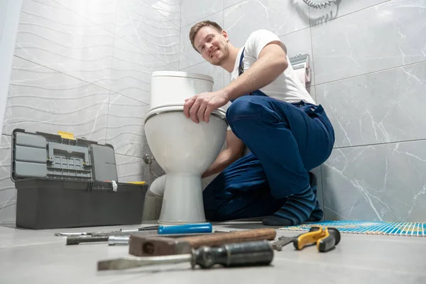Sonriente hombre fontanero en uniforme reparación inodoro utilizando kit de instrumentos se ve feliz servicio de reparación profesional — Foto de Stock