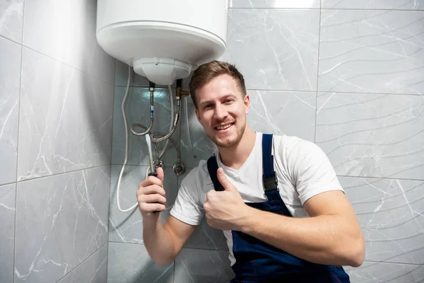Sonriente hombre trabajador en uniforme con destornillador en una mano y mostrando como con otra mano reparación de caldera en casa en servicio de reparación profesional toilette — Foto de Stock