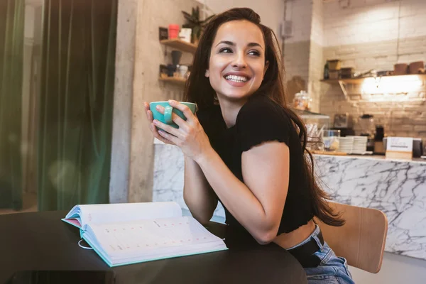 Joven hermosa morena sonriente mujer bebe café caliente en el café se ve feliz mientras trabaja en su planificador — Foto de Stock