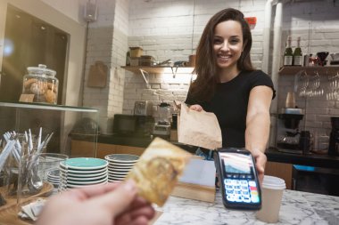 Barın arkasında elinde sıcak kahve bardağıyla duran barmen kadın ve yemek çantası ödeme terminalini almak için.