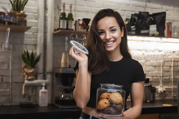 young smiling beautiful woman barista enjoying nice aroma while opening jar with cookies lokking happy