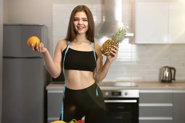 Ung vältränad kvinna med centimeter rund hals klädd i svart topp och svarta leggings med ananas i ena handen och orange i en annan dietologi och näring — Stockfoto