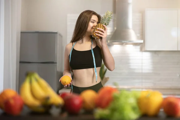 Ung vältränad kvinna med centimeter rund hals bär svart topp och leggings luktar ananas stående i köket full av frukt dietologi och näring — Stockfoto
