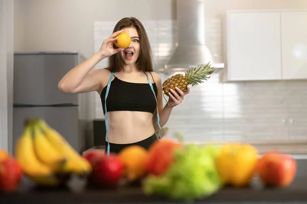 Jovem mulher apto com centímetro de pescoço redondo vestindo top preto e leggings mantém laranja perto de seu olho em pé na cozinha cheia de dietologia de frutas e nutrição — Fotografia de Stock
