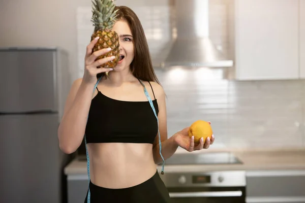 Mujer joven en forma con cuello redondo centímetro usando top negro y polainas sostiene piña cerca de su cara de pie en la cocina dietología y nutrición — Foto de Stock