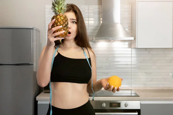 Giovane donna in forma con collo rotondo centimetri indossando top nero e leggings tiene ananas vicino al viso in piedi in cucina dietologia e nutrizione cercando sorpreso — Foto Stock