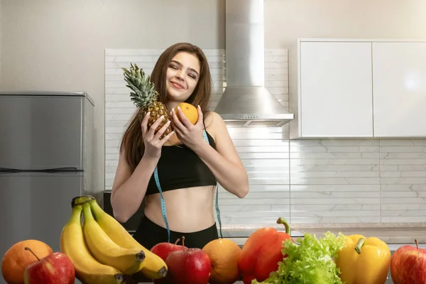 Žena s centimetrem kulatý krk na sobě černý top a legíny objetí ananas a pomeranč v kuchyni plné ovoce dietologie a výživy — Stock fotografie