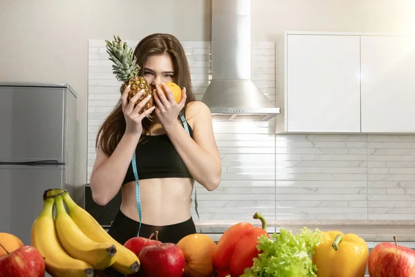 Žena s centimetrem kulatý krk nosí černý top a kamaše kousne ananas a pomeranč v kuchyni plné ovoce dietologie a výživy — Stock fotografie
