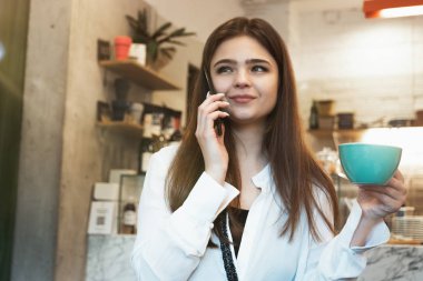 Genç, güzel, esmer, gülümseyen kadın kafede sıcak kahve içiyor. Akıllı telefonuyla konuşurken mutlu görünüyor.