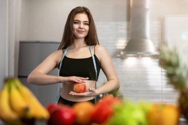 İki elinde de elma tutan yuvarlak boyunlu kadın mutfaktaki meyvelerle birlikte beslenme ve beslenme düzenine bakıyor.
