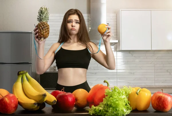 Mulher com centímetro de pescoço redondo com abacaxi em uma mão e laranja em outra de pé na cozinha cheia de frutas olhando dietologia decepcionada e nutrição — Fotografia de Stock