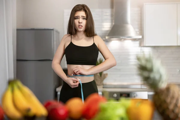 Mulher com centímetro em volta da cintura vestindo top preto e leggings de pé na cozinha cheia de frutas olhando chateado dietologia e nutrição — Fotografia de Stock