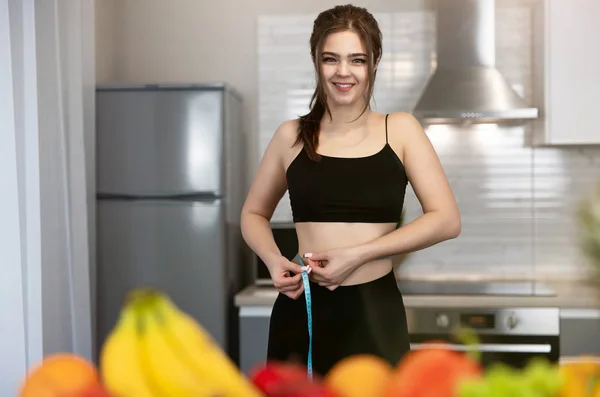 woman with centimeter round her waist checking parameters wearing black top and leggings standing in kitchen full of fruits looking satisfied dietology and nutrition