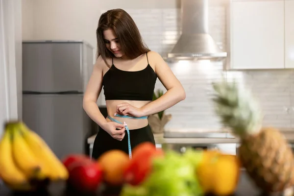 Mulher com centímetro em volta da cintura verificando parâmetros vestindo top preto e leggings de pé na cozinha cheia de dietologia frutas e nutrição — Fotografia de Stock