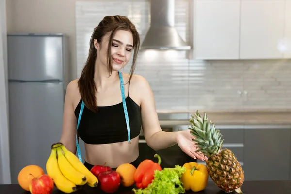 Jovem mulher apto com centímetro de pescoço redondo vestindo top preto e leggings de pé na cozinha cheia de frutas toca abacaxi, dietologia e nutrição — Fotografia de Stock