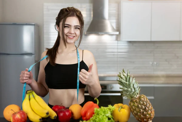 Jovem mulher apto com centímetro de pescoço redondo vestindo top preto e leggings de pé na cozinha cheia de frutas mostrando como dietologia sinal e nutrição — Fotografia de Stock