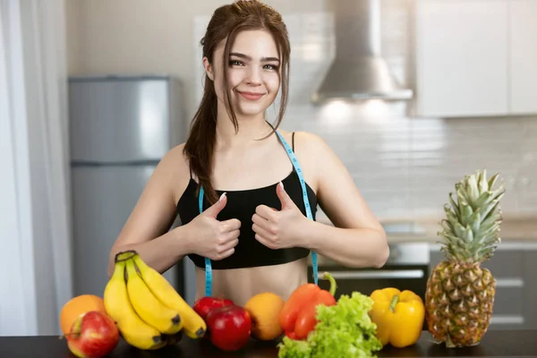 Fit woman with centimeter round neck wearing black top standing in the kitchen full of fruits showing like sign with both hands dietology and nutrition — Stock Photo, Image