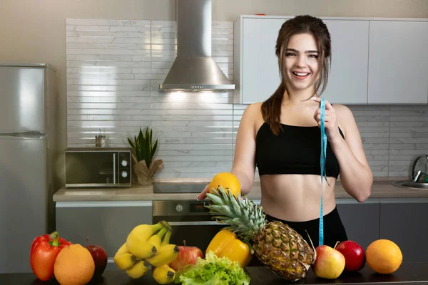 Mulher apto com centímetro em uma mão e laranja em outra vestindo top de esportes preto em pé na cozinha cheia de frutas, dietologia e nutrição — Fotografia de Stock