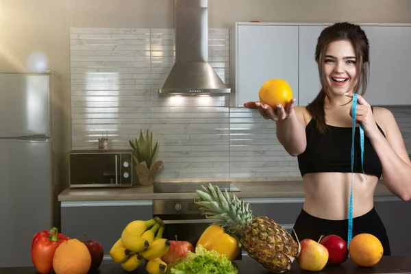 Mulher apto segurando centímetro em uma mão e laranja em outra vestindo top de esportes preto em pé na cozinha cheia de frutas, dietologia e nutrição — Fotografia de Stock