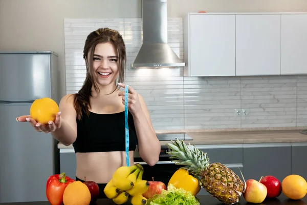 Mulher apto com centímetro em uma mão esticando laranja vestindo top de esportes preto em pé na cozinha cheia de frutas, dietologia e nutrição — Fotografia de Stock