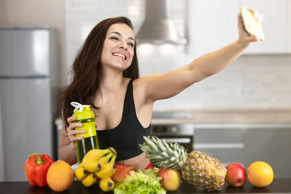Fit young woman wearing sexy black sports top shooting selfie standing in the kitchen full of fruits, dietology and nutrition — Stock Photo, Image