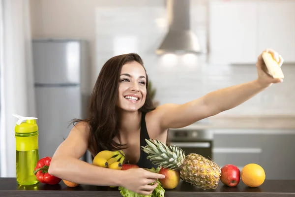 Mujer joven en forma usando sexy negro deportes top tiro selfie en su teléfono inteligente de pie en la cocina llena de frutas, dietología y nutrición — Foto de Stock