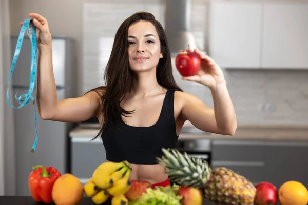 Fit žena s centimetrem v jedné ruce a jablko v druhé v černé sportovní top stojící v kuchyni plné ovoce, dietologie a výživy — Stock fotografie