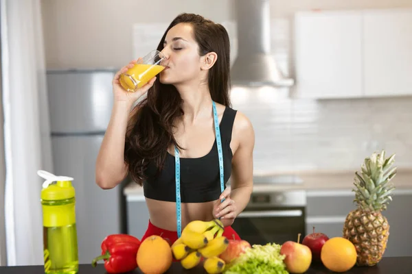Fit žena s centimetrem kulatý krk na sobě černé sportovní top stojící v kuchyni plné ovoce nápoje pomerančový džus ze skleněné dietologie a výživy — Stock fotografie