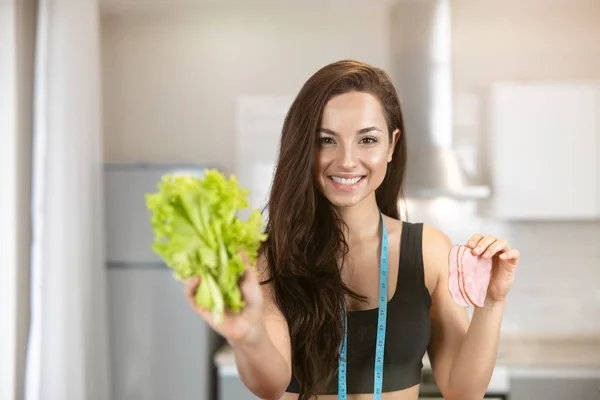 Joven mujer en forma con centímetro alrededor de su cuello sosteniendo ensalada fresca en una mano y rebanada de jamón en otra se ve feliz, nutrición y dietología — Foto de Stock