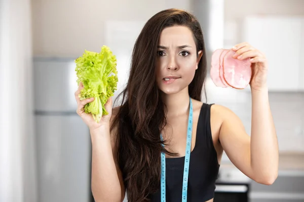Fiatal fitt nő centiméterrel a nyakában tartja friss saláta az egyik kezében, és szelet sonka a másikban úgy néz ki, tétova, táplálkozás és dietológia — Stock Fotó