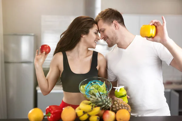 fit couple man and woman leaning to each other in kitchen full of fruits and vegetables, he holds pepper, she holds apple, nutrition and dietology