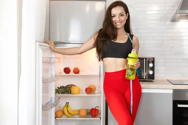 Passform kvinna med centimeter runt halsen bär sportkläder stående i köket nära öppen kyl full av frukt och grönsaker, dietologi och näring — Stockfoto