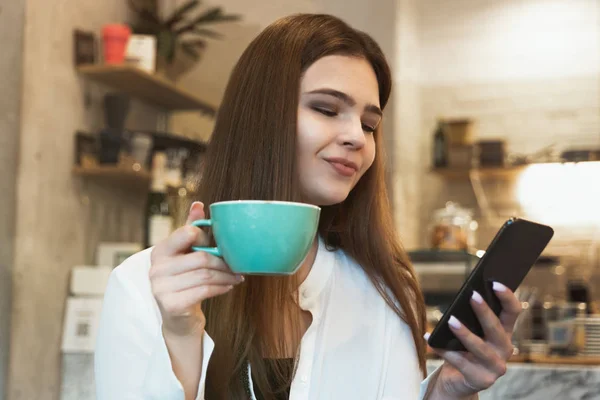 Joven hermosa morena sonriente mujer bebe café caliente en el café se ve feliz de comprobar su teléfono inteligente — Foto de Stock