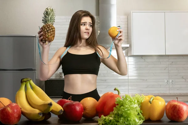Žena s centimetrem kulatého krku s ananasem v jedné ruce a pomerančem v druhé stojící v kuchyni plné ovoce vypadající zmateně dietologie a výživy — Stock fotografie