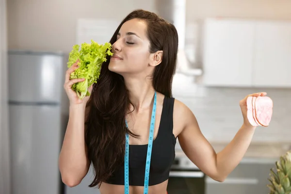 Joven mujer en forma con centímetro alrededor de su cuello sosteniendo ensalada fresca en una mano y rebanada de jamón en otro se ve hambriento, nutrición y dietología — Foto de Stock