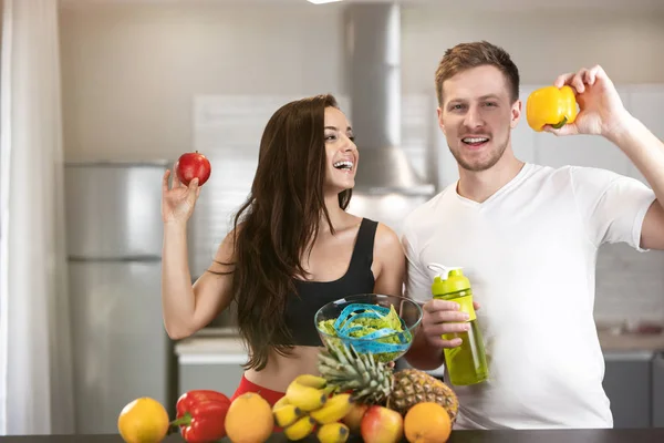 Jong fit lachend paar man en vrouw staan in de keuken vol met groenten en fruit, hij houdt peper, ze houdt appel, voeding en voedingsleer — Stockfoto