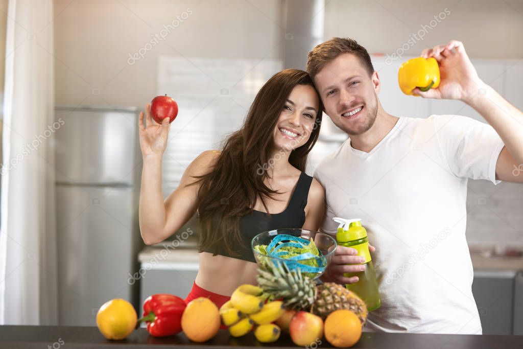 young happy fit couple man and woman standing in the kitchen full of fruits and vegetables, he holds pepper, she holds apple, nutrition and dietology