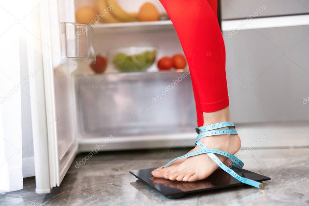 woman's legs wrapped in centimeter standing on scales to measure weight in the kitchen near open fridge full of fruits and vegetables, dietology and nutrition