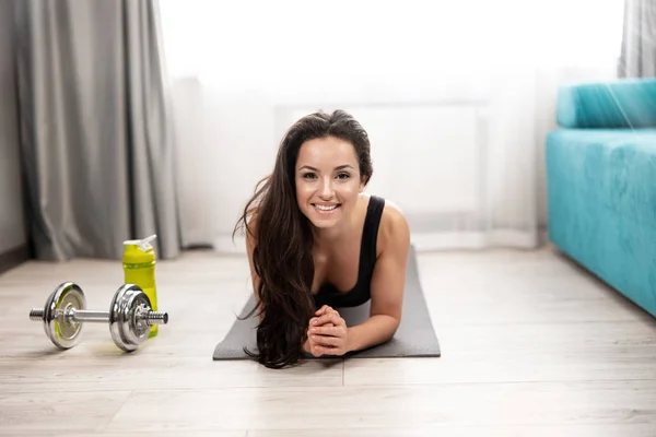 smiling fit brunette woman wearing sportwear standing in plank while doing everyday training exercises with dumbbell on the floor at home, sportive lifestyle