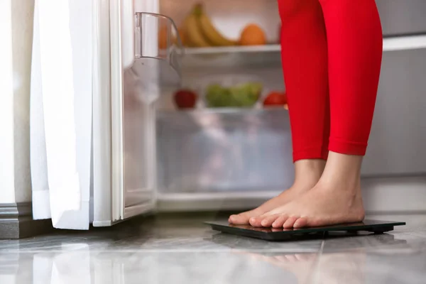 woman\'s legs standing on scales to measure weight in the kitchen near open fridge full of fruits and vegetables, dietology and nutrition