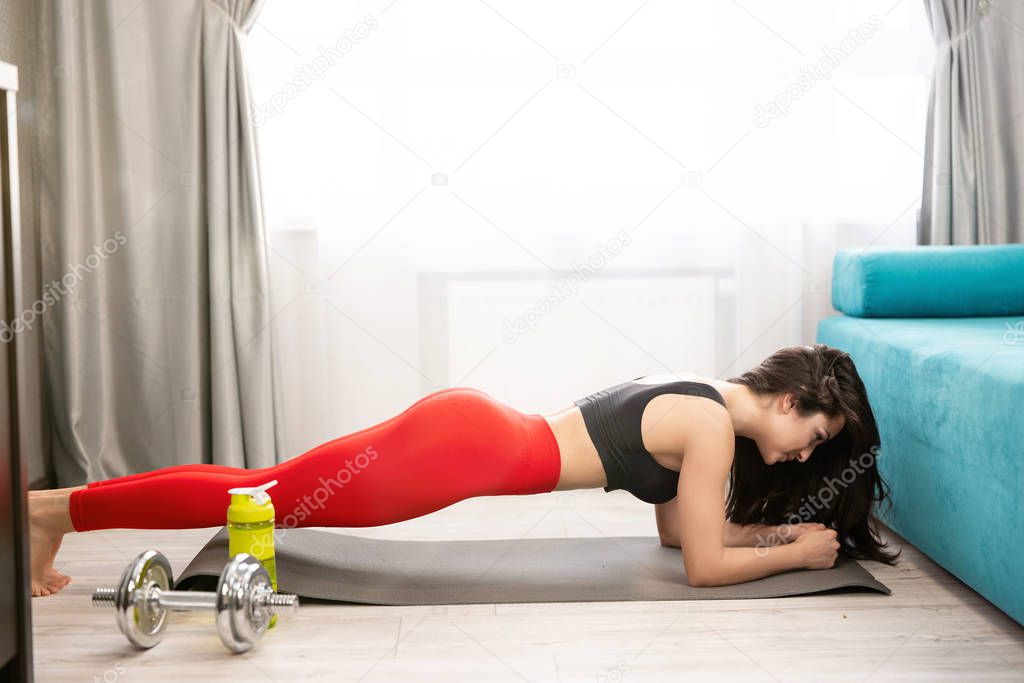 young fit brunette woman wearing sportwear standing in plank after doing everyday warm up exercises with dumbbell on the floor at home, sportive lifestyle