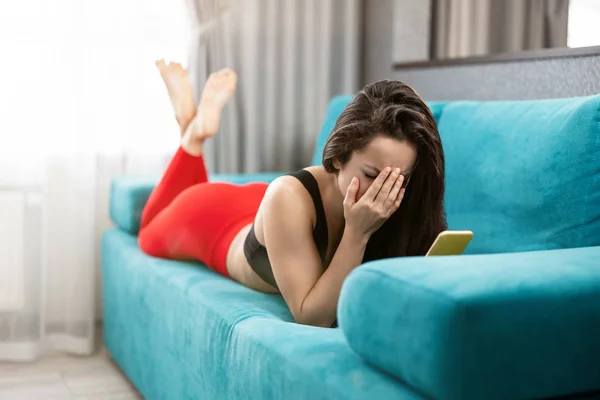 Fit brunette woman in red sport leggings and black top lying on the sofa looking upset after reading unpleasant news in her smartphone, melancholic mood — Stock Photo, Image