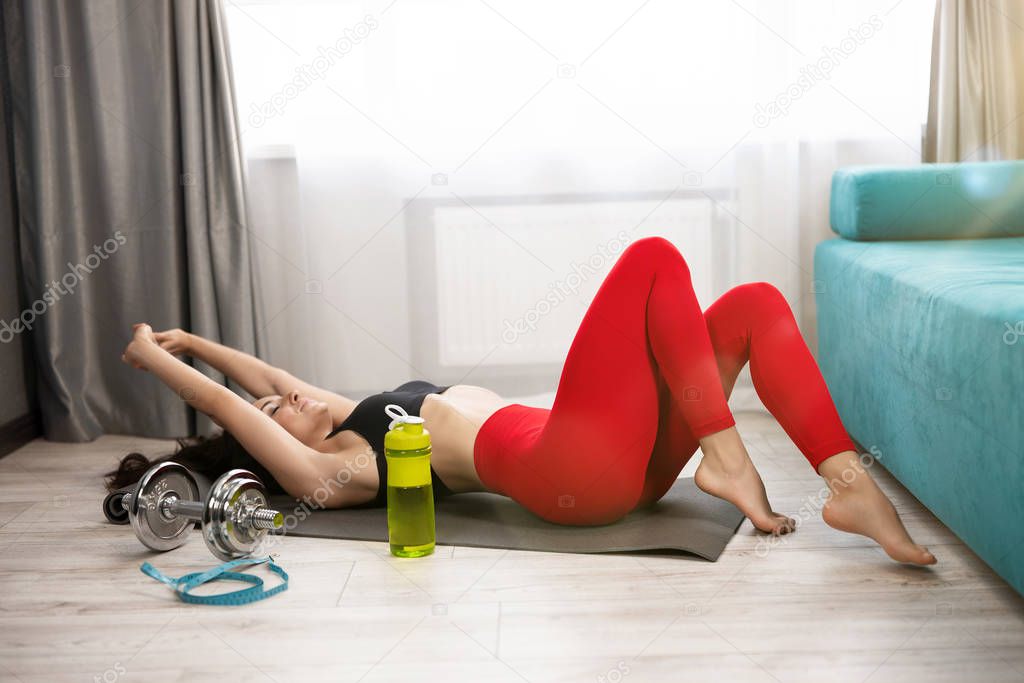 young fit brunette woman in sportwear lies on the floor looking tired after daily home workout on the mat in living room, sportive lifestyle