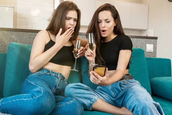 Deux jeunes belles femmes amies buvant du champagne sur le canapé regardant choqué et surpris en regardant smartphone, célébrer l'amitié — Photo