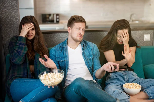 Tre unge venner to vakre kvinner og en kjekk mann med fjernkontroll som ser på dramafilm på sofaen og spiser popkorn, jenter som reagerer følelsesmessig, vennskap – stockfoto