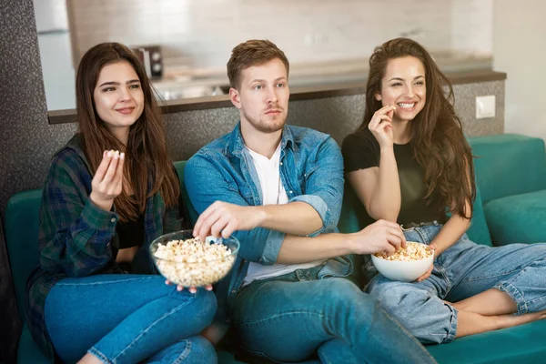 Trois jeunes amis deux belles femmes et un bel homme regardant la télévision sur le canapé manger du maïs pop, amitié — Photo