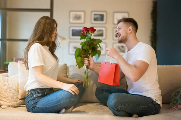 Unga par sitter på soffan, man överraskande fru med blommor och gåva på helgonet Alla hjärtans dag, glad årsdag — Stockfoto